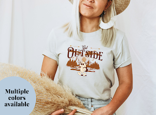 Woman wearing a shirt that says Get Outside with a graphic of a groovy dog hiking with a backpack in the mountains