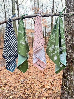 Four bandanas hanging on a tree branch. There are two green bandanas, one beige bandana and a black and white bandana.