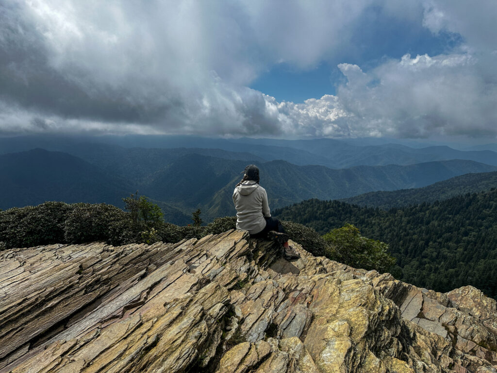 A woman with her back to the camera sitting on a cliff looking at a cloudy sky over the smoky mountains from mount leconte