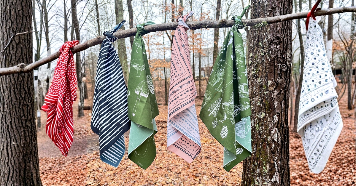 Six bandanas hanging on a tree branch. There are two green bandanas, one beige bandana, a red one, white one and a black and white bandana.