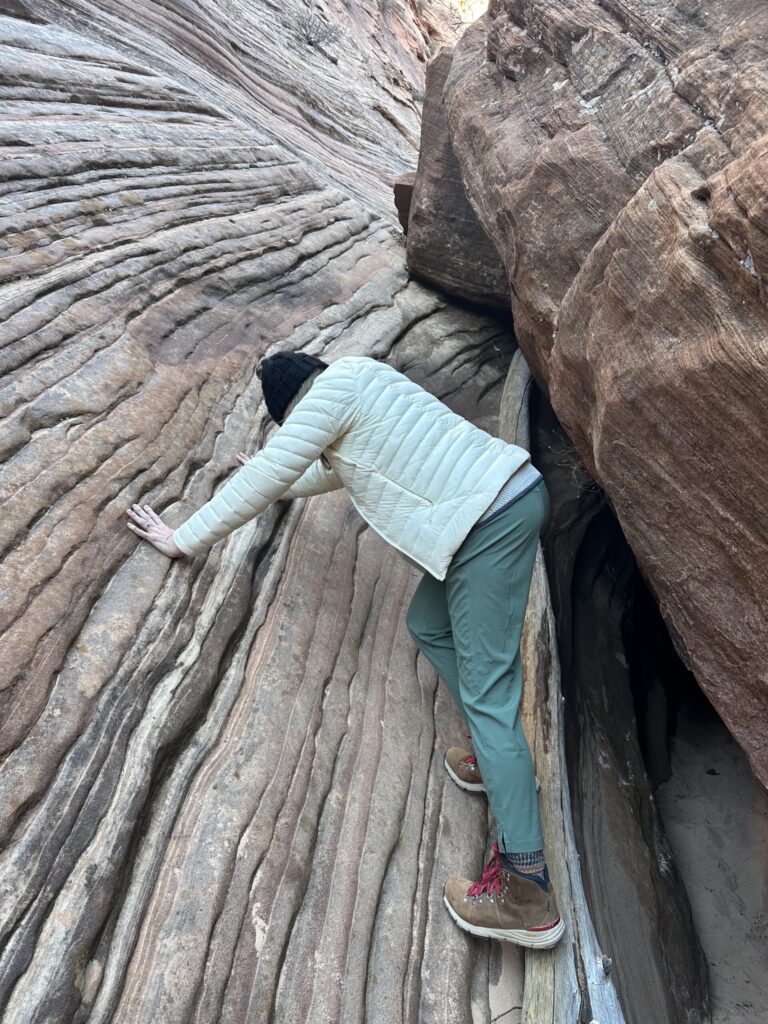 A woman in a white jacket and green pants pushing against a rock wall
